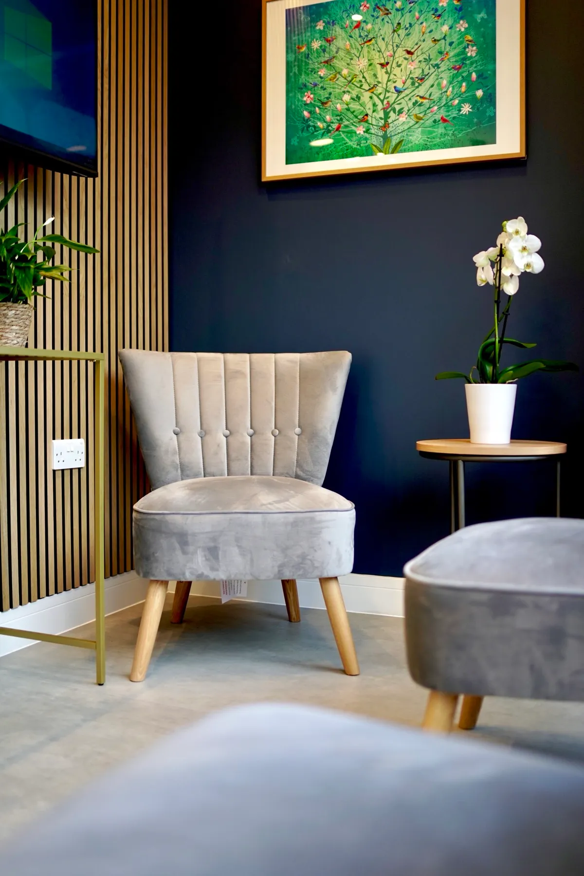 Modern waiting room corner with a grey velvet chair, wooden legs, a small table with a white orchid, and a colourful tree painting on a dark blue wall.