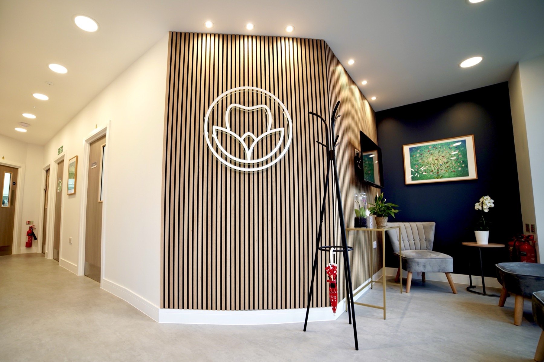 Modern waiting area with wooden slat wall featuring a tooth logo, gray chairs, a coat rack with a red umbrella, plants, and framed artwork.