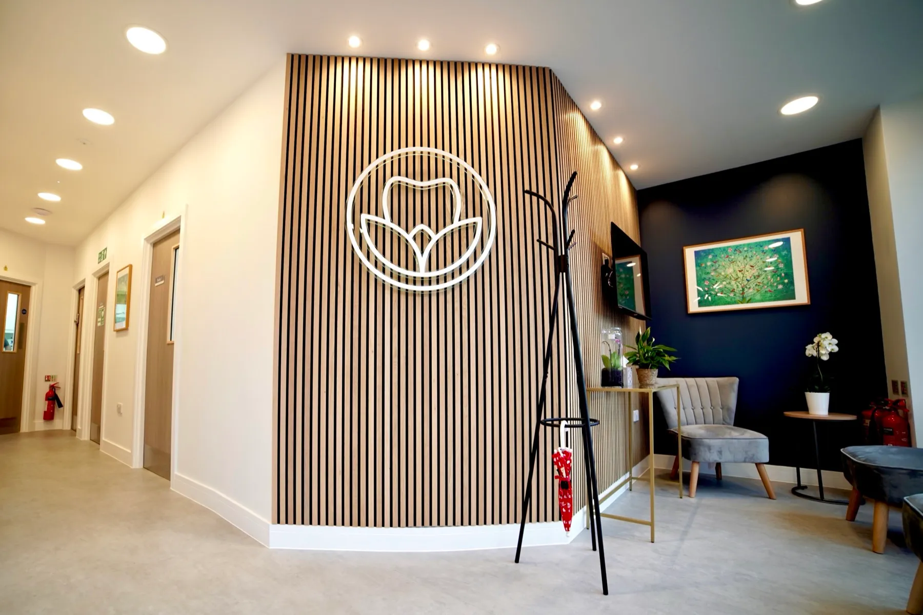Modern waiting area with wooden slat wall featuring a tooth logo, gray chairs, a coat rack with a red umbrella, plants, and framed artwork.