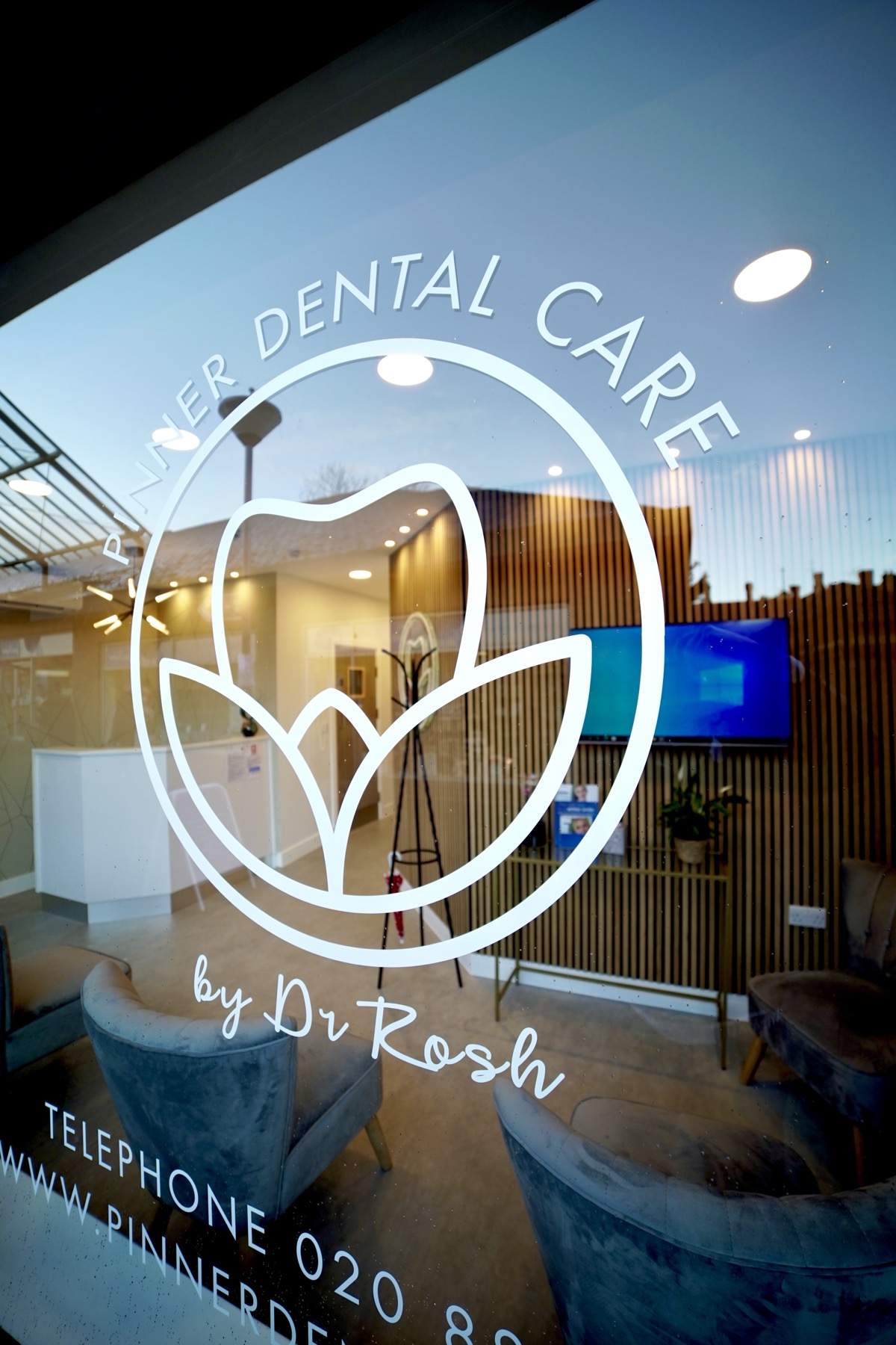 Reception area of Pinner Dental Care with chairs and wooden slat wall visible through the glass door featuring the dental clinic logo and contact information.