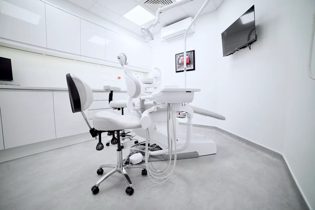 Modern dental clinic room with white walls, dental chair and equipment, a dental stool, white cabinets, a flat-screen TV mounted on the wall, and a framed picture.