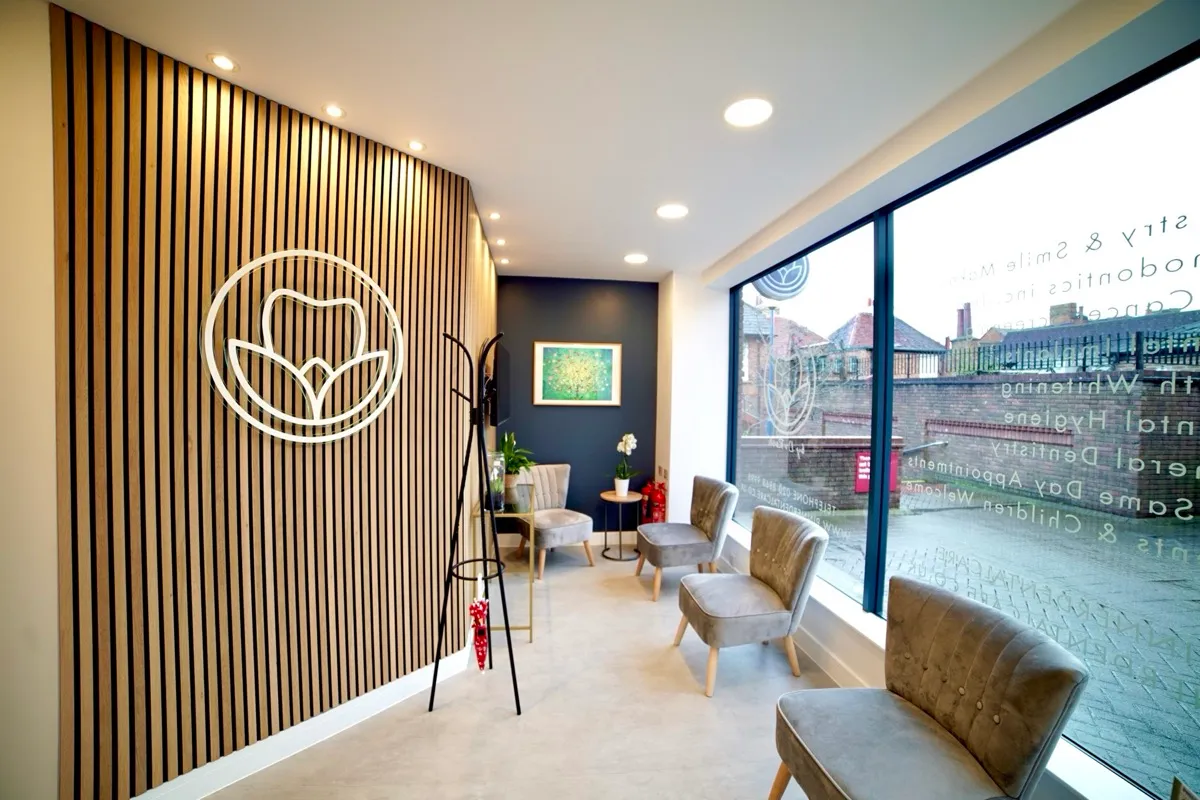 Modern waiting area with wooden slat wall featuring tooth and leaf logo, gray upholstered chairs along large window, coat rack, indoor plants, and framed artwork on a dark blue accent wall.