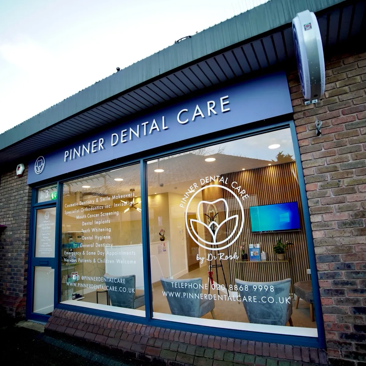 Exterior view of Pinner Dental Care with large windows displaying services like cosmetic dentistry, orthodontics, dental implants, and emergency appointments, featuring a modern waiting area with chairs and a wall-mounted TV inside.