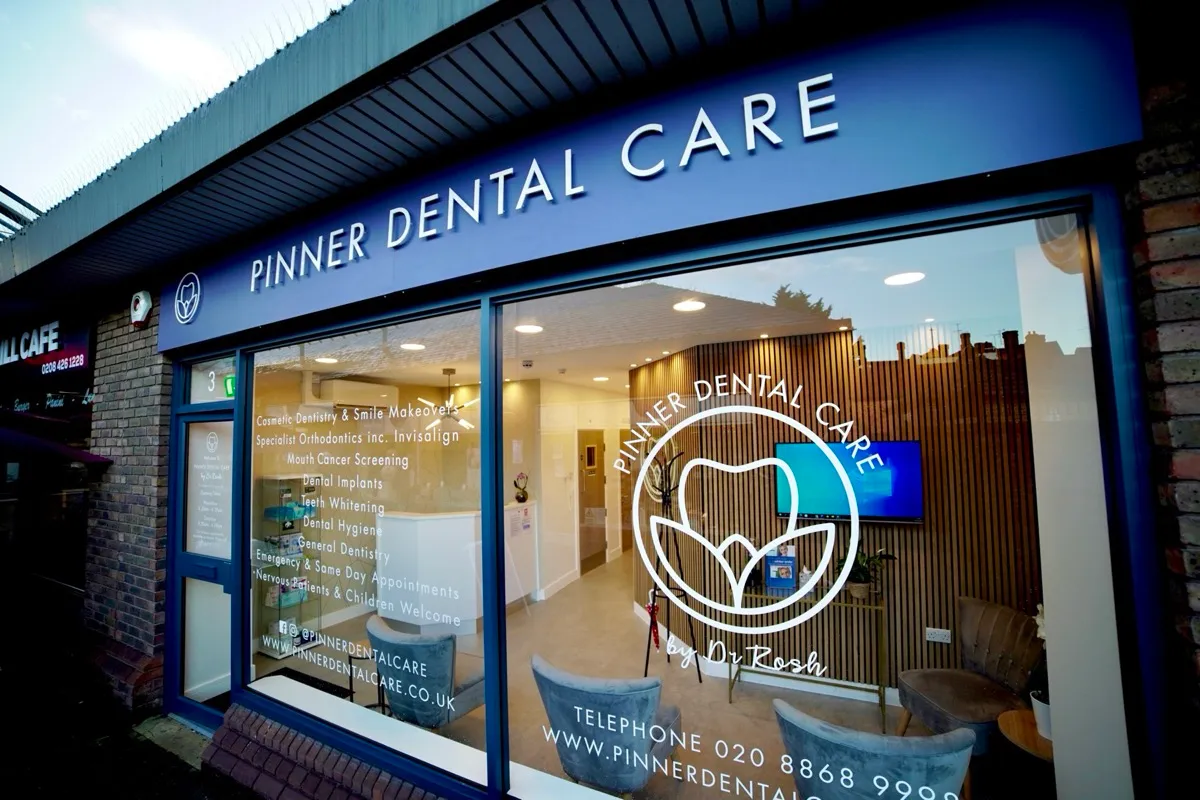 Front view of Pinner Dental Care clinic showing its blue signage with white letters, a large window displaying the clinic's logo, services like cosmetic dentistry, orthodontics, dental implants, teeth whitening listed on the glass, and a glimpse of the modern waiting area inside with chairs and a TV screen.