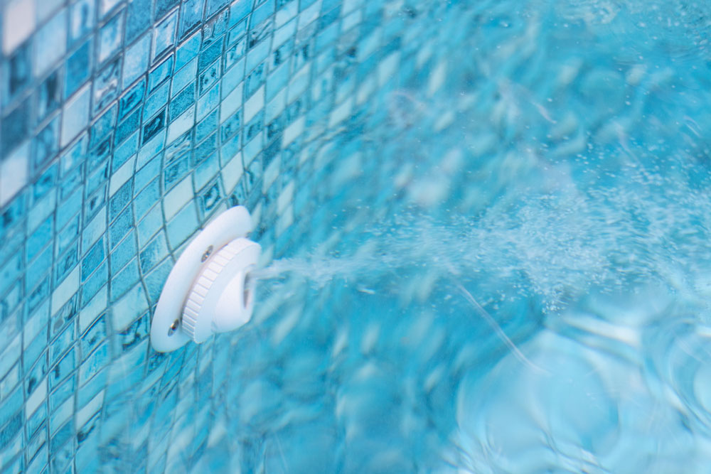 Pool return fitting in clear blue water beside a tiled pool wall