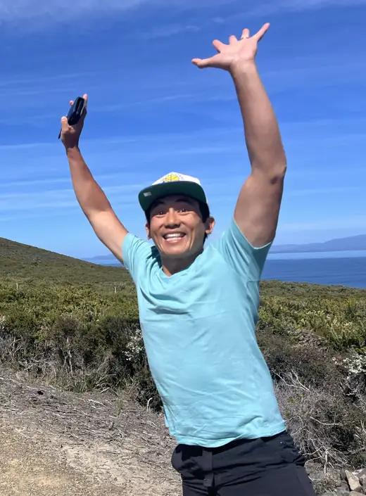 Benn Lim with his arms in the air, grinning on a coastal walk near a lighthouse