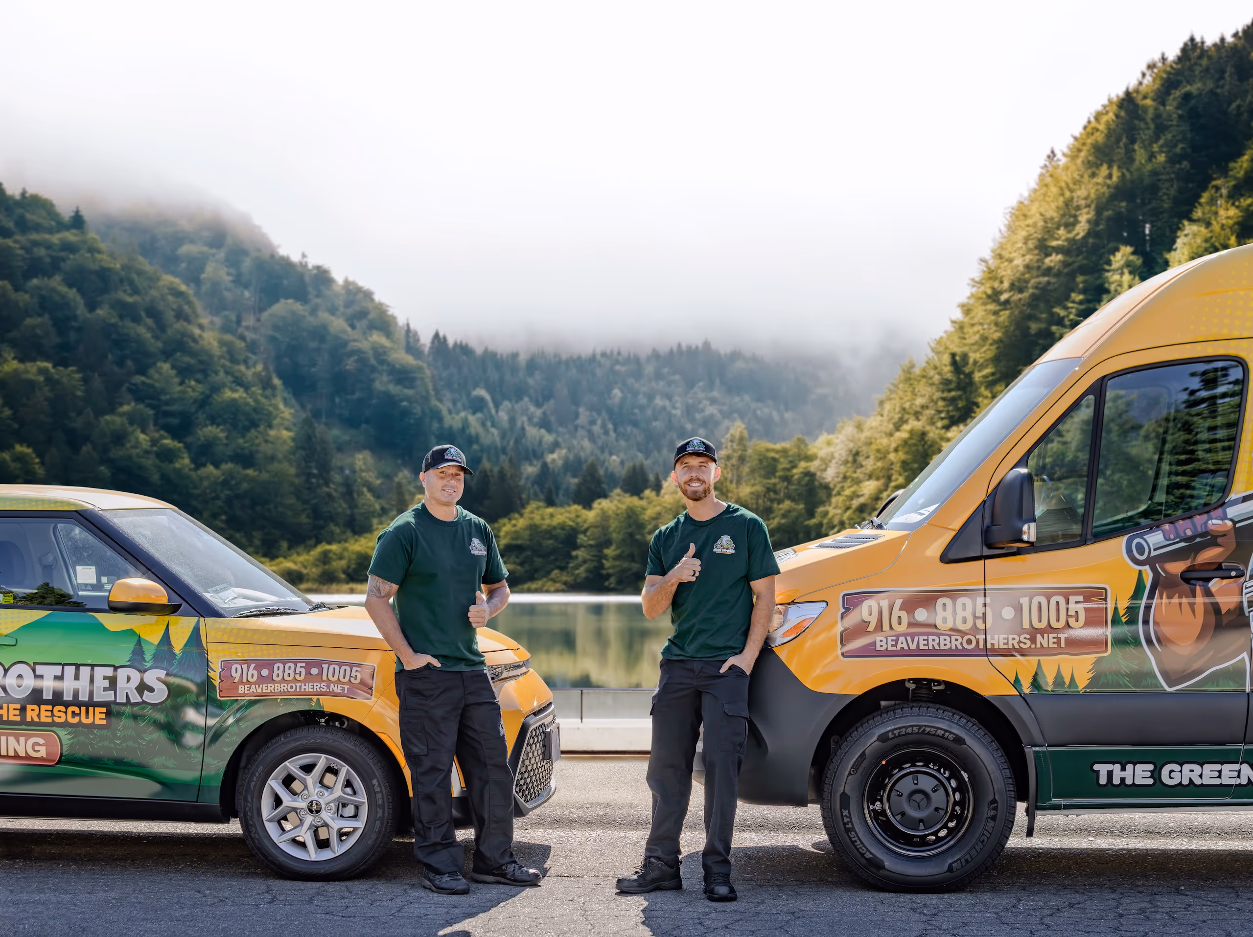Two men in matching green shirts and black pants giving thumbs up while standing between two branded vehicles by a lake with forested hills in the background.