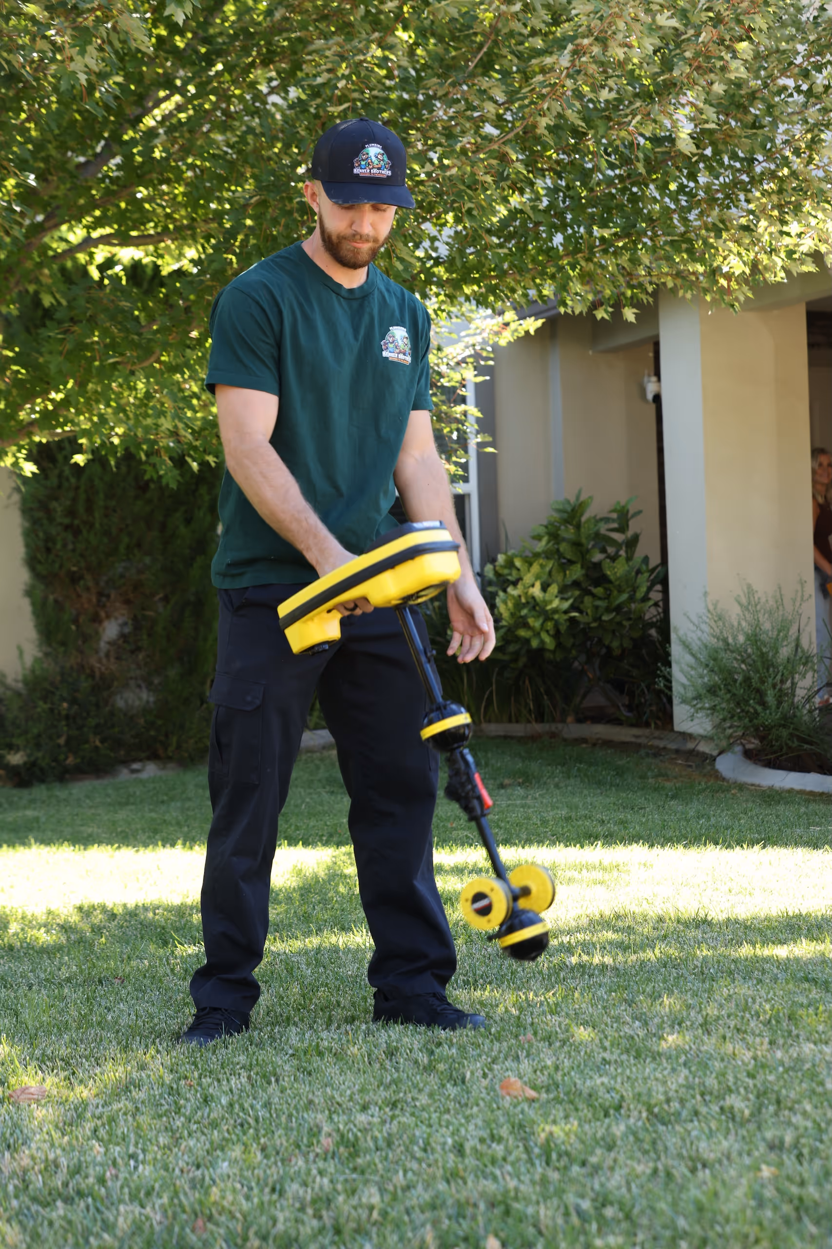 Beaver Brothers plumber in uniform using a yellow pipe and utility locator tool on a residential lawn.