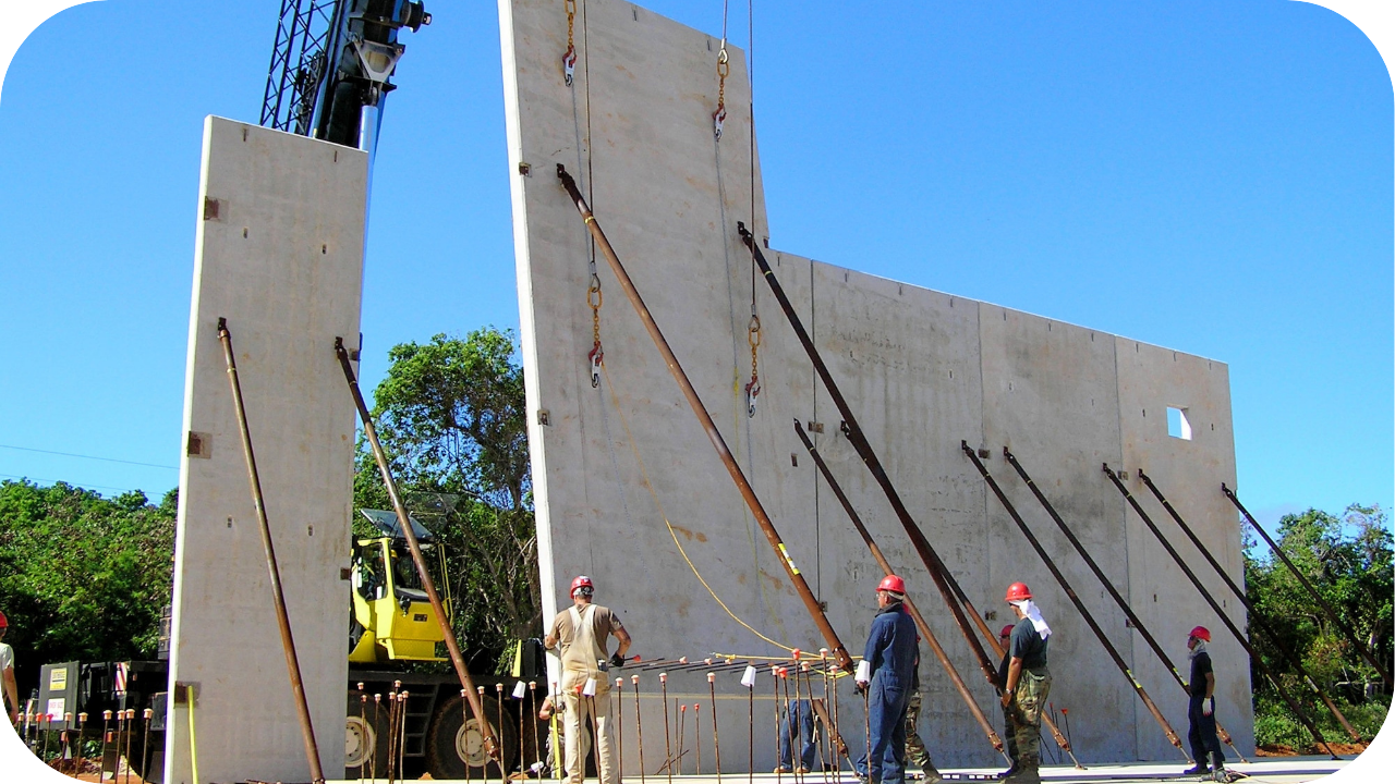 A construction crew uses a crane to install massive, precast concrete wall panels, securing them with long metal braces.
