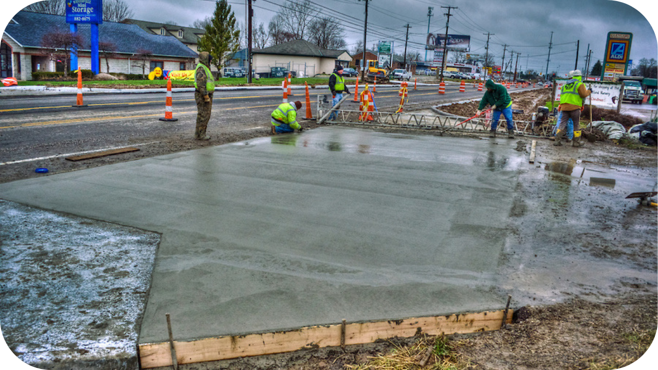 A crew of workers finishes a large, new wet concrete slab next to a road on an overcast day.