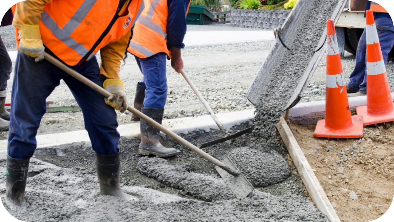 Two workers in orange safety vests use shovels to spread wet concrete as it flows from a truck chute into a formwork.