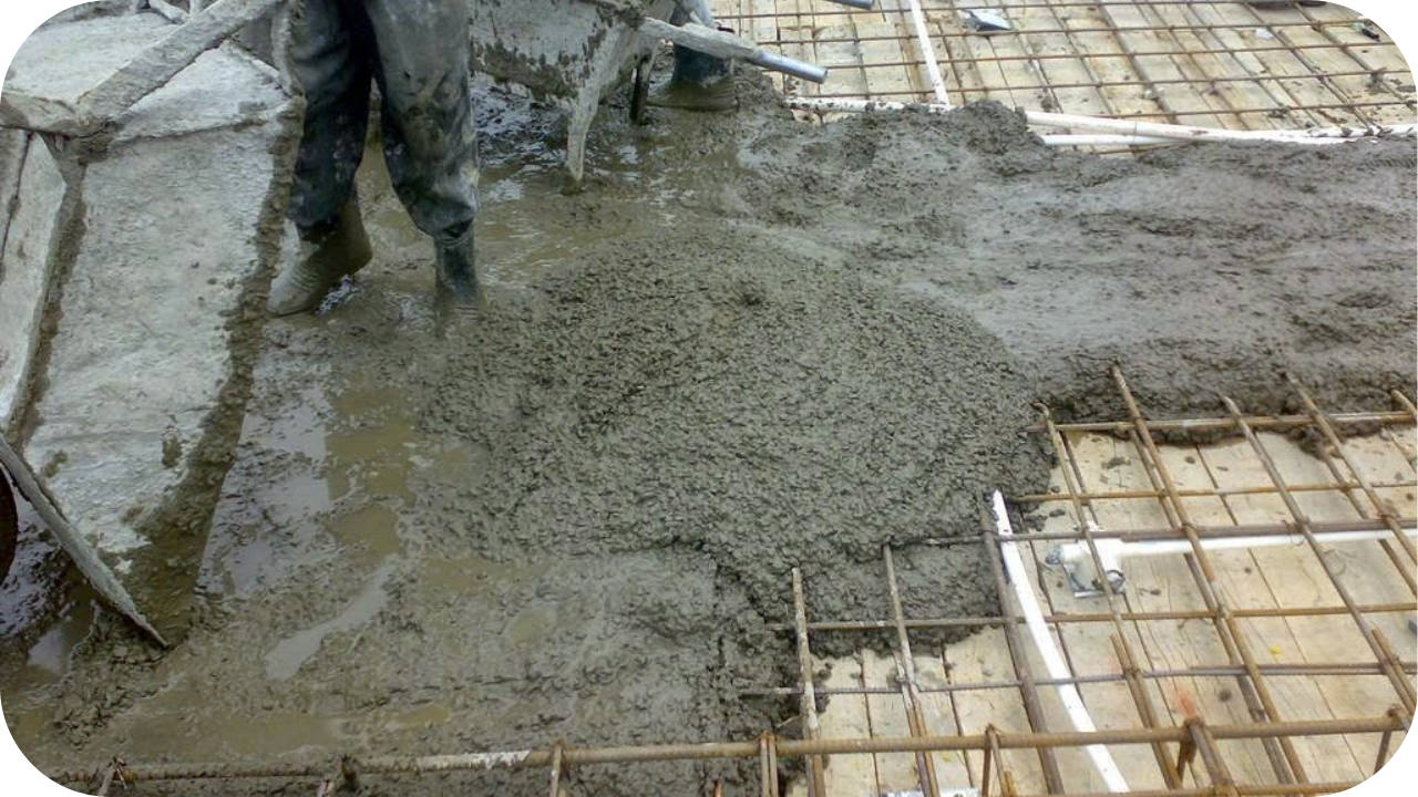 Wet concrete is poured from a wheelbarrow onto a muddy construction site with a steel rebar grid.
