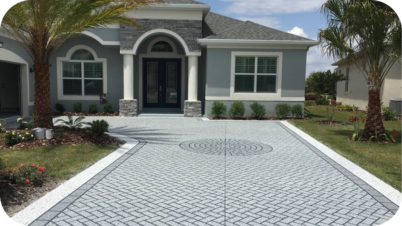 A decorative concrete driveway with a grey herringbone paver pattern and a circular medallion, in front of a modern grey house.