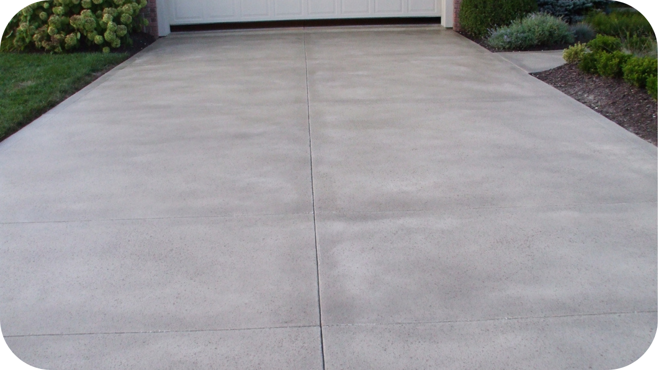 A clean, smooth concrete driveway with control joints, leading up to a white garage door and bordered by a green lawn.