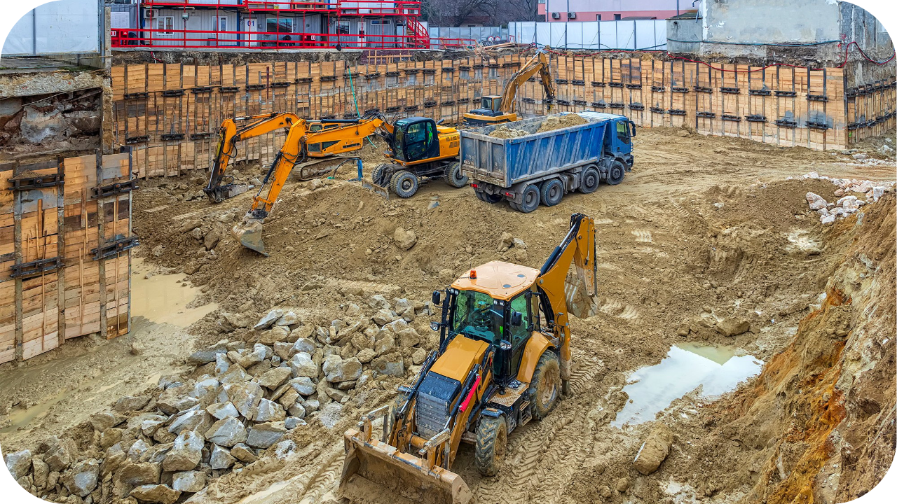 A deep excavation pit on a construction site, with two excavators and a large dump truck working in the mud below.