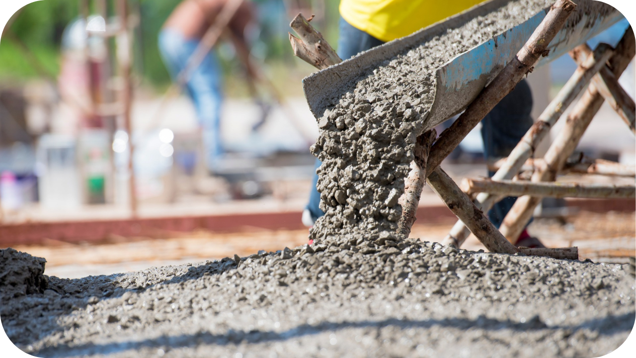 A low-angle view of wet concrete flowing from a metal wheelbarrow chute onto a pile on the ground at a construction site.