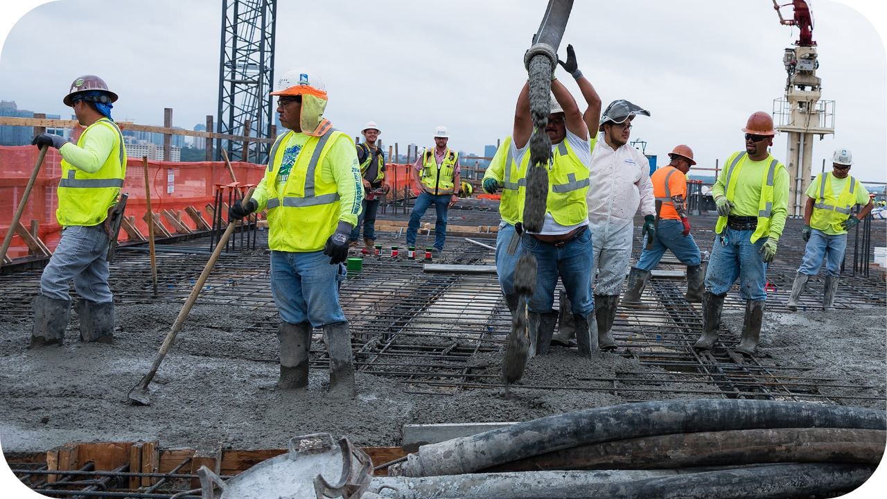 A large crew of construction workers in hard hats pours the concrete slab on the top floor of a high-rise building.