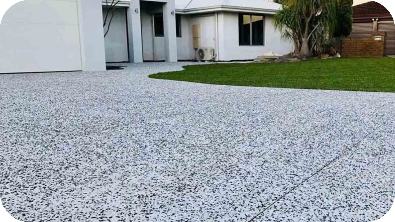 A residential driveway with a light grey epoxy flake coating, in front of a modern white house and a green lawn.