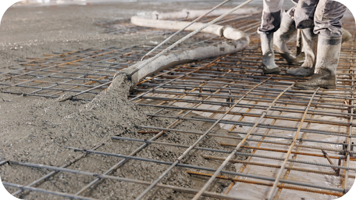 Workers in boots guiding concrete pump hose to pour fresh concrete over steel reinforcement mesh on a building site.