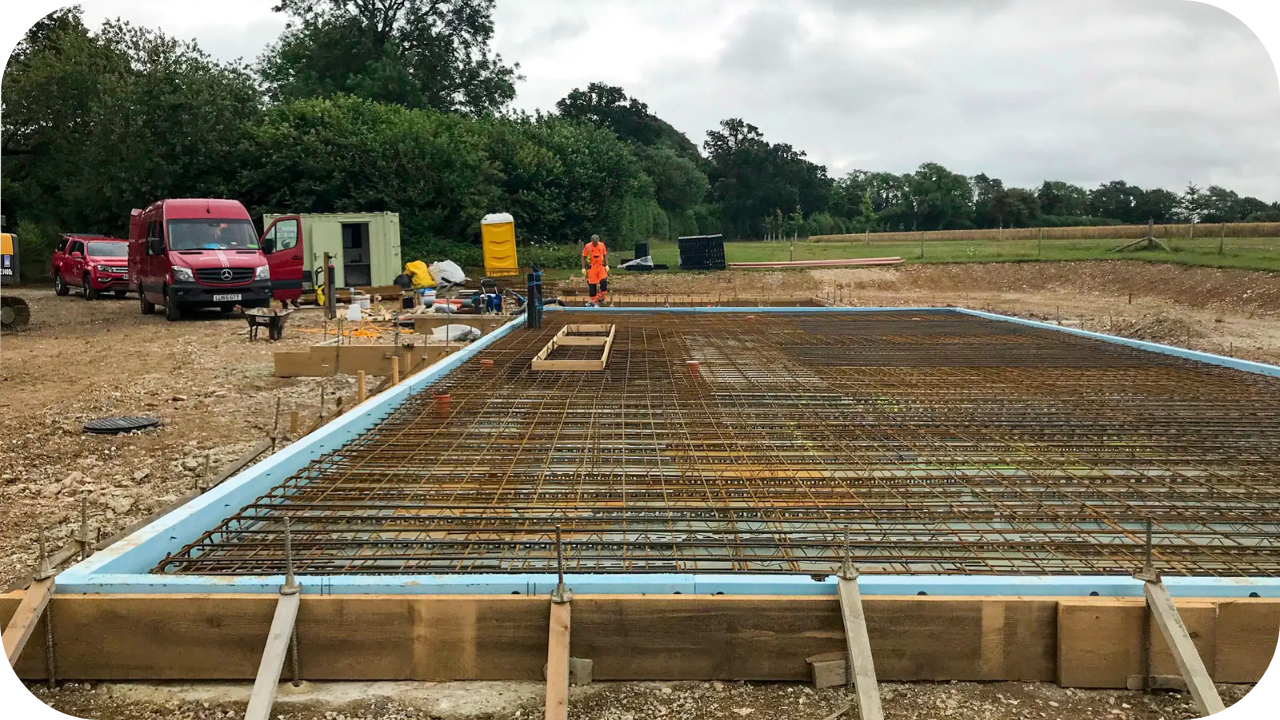 Construction site prepared with rebar grid and timber formwork before concrete pour, with workers and vehicles nearby.