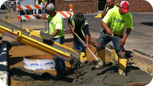A crew of three workers in yellow shirts pours and spreads wet concrete from a long chute to form a new curb or sidewalk.