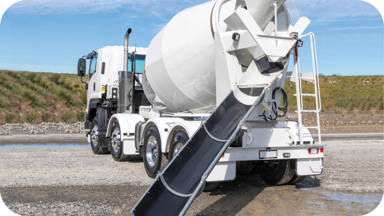 A white Urban Pumping truck is parked on a gravel construction site, positioned to receive concrete from a larger mixer truck.