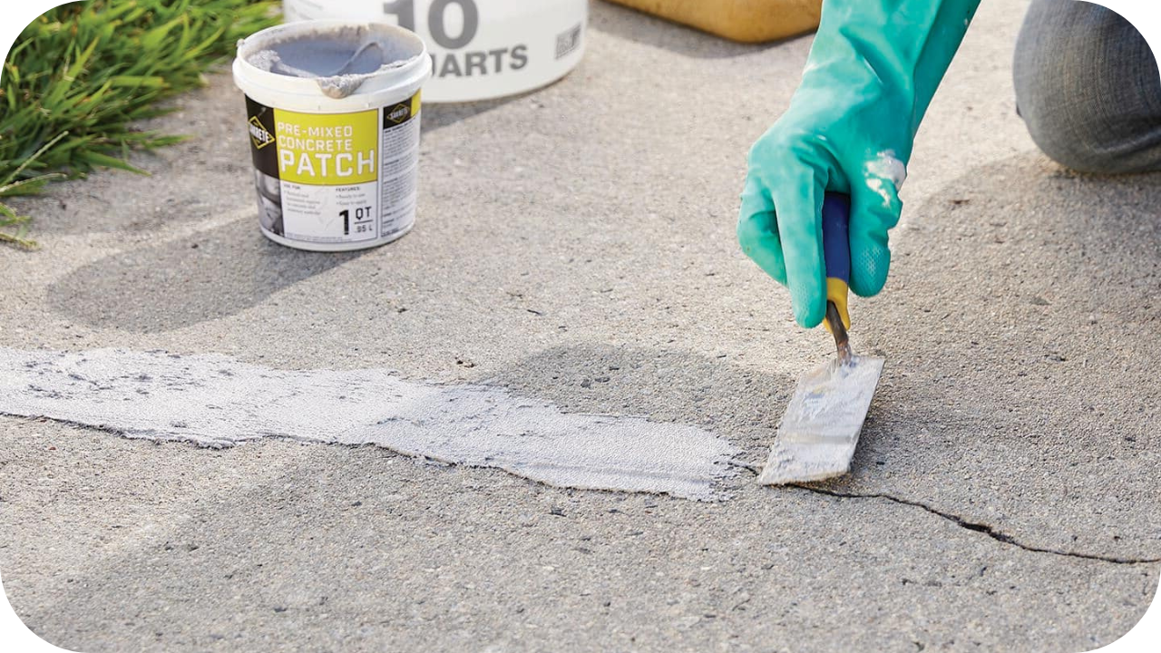 Hand in green glove applying premixed concrete patch to repair a crack on a paved surface.