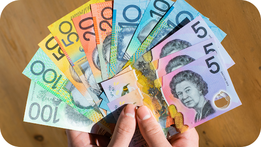 Hand holding a fan of Australian dollar banknotes in denominations of 5, 10, 20, 50, and 100.