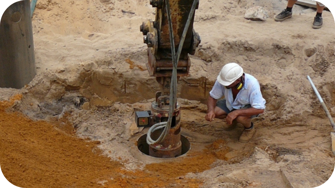 Worker inspecting auger drill during bored pier foundation installation, ensuring correct depth and alignment on sandy site.