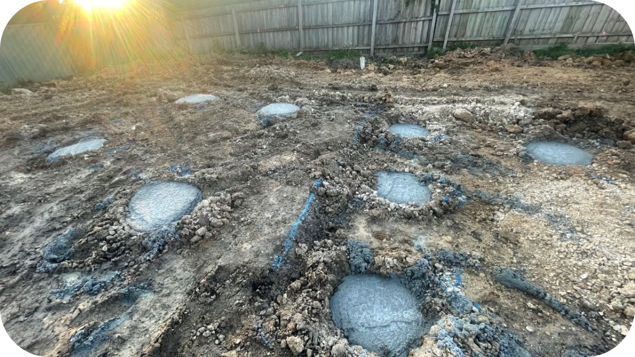 Freshly poured concrete bored piers visible in marked excavated holes on residential construction site at sunset.