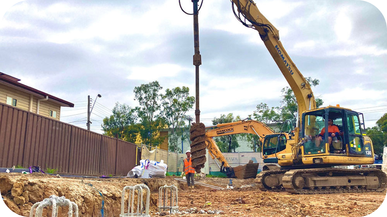 Excavator with auger attachment drilling deep bored pier foundations on construction site with workers overseeing process.
