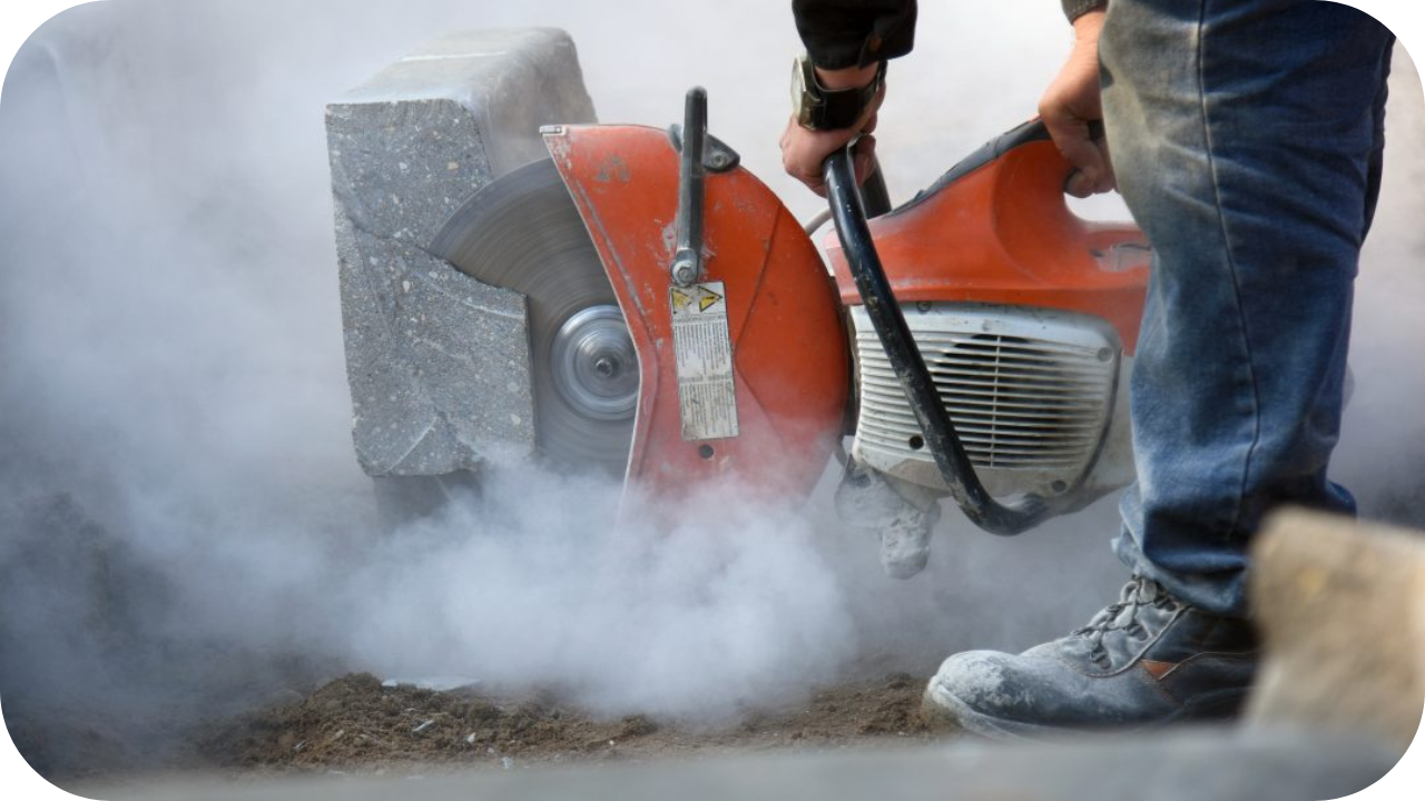 A worker uses a large, orange handheld saw to cut a concrete block, creating a thick cloud of white dust.