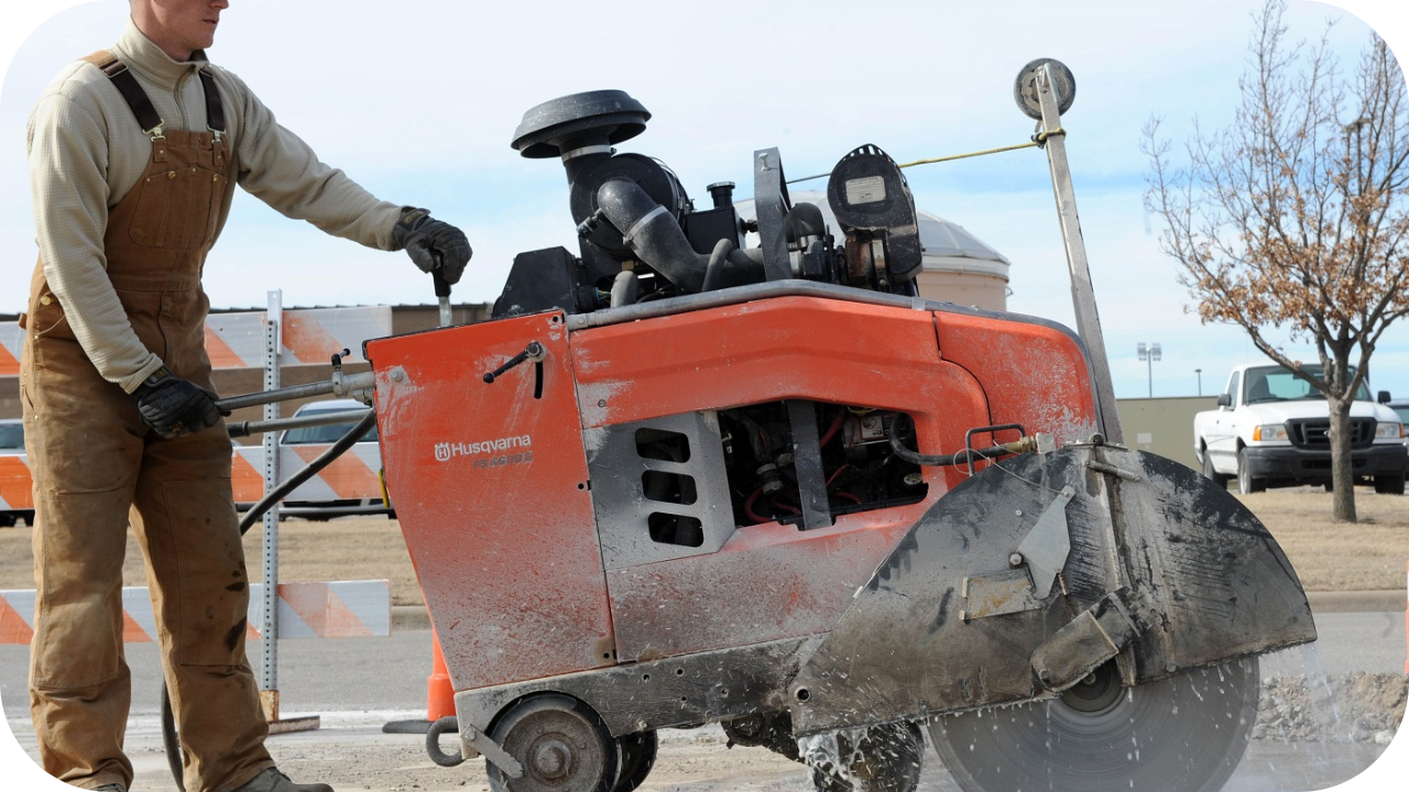 A worker in overalls operates a large, orange walk-behind floor saw to cut a line in an asphalt road.