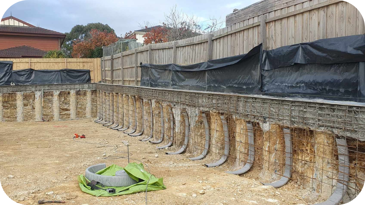 A deep residential excavation for a basement or pool, with the earth walls stabilized by concrete piers, rebar mesh, and drainage pipes.