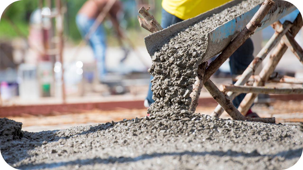 A low-angle view of wet concrete flowing from a metal wheelbarrow chute onto a pile on the ground at a construction site.