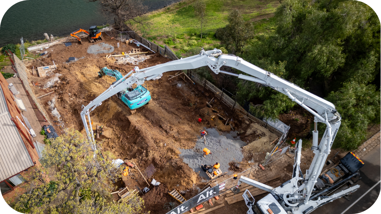 Overhead view of a construction site showing concrete pumping machinery and workers preparing the foundation near a waterway.