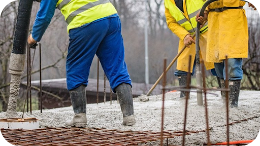 Construction workers in rain gear pouring concrete over rebar on a wet building site.