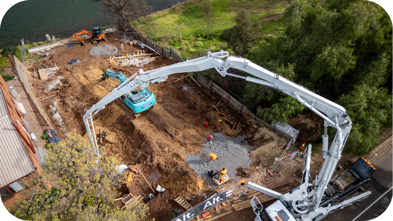 Aerial view of a residential construction site beside a river, showing a white boom pump truck pouring concrete.