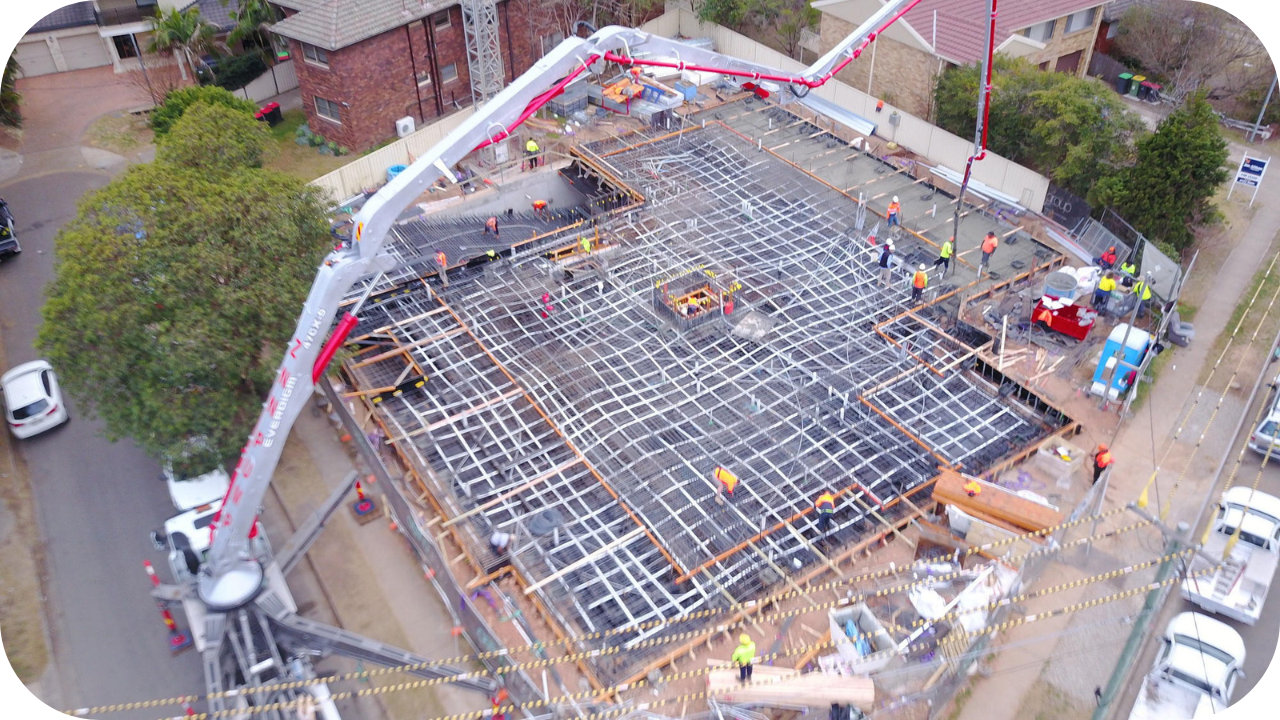Aerial view of workers and concrete pumps building a post-tensioned slab foundation at an urban construction site.