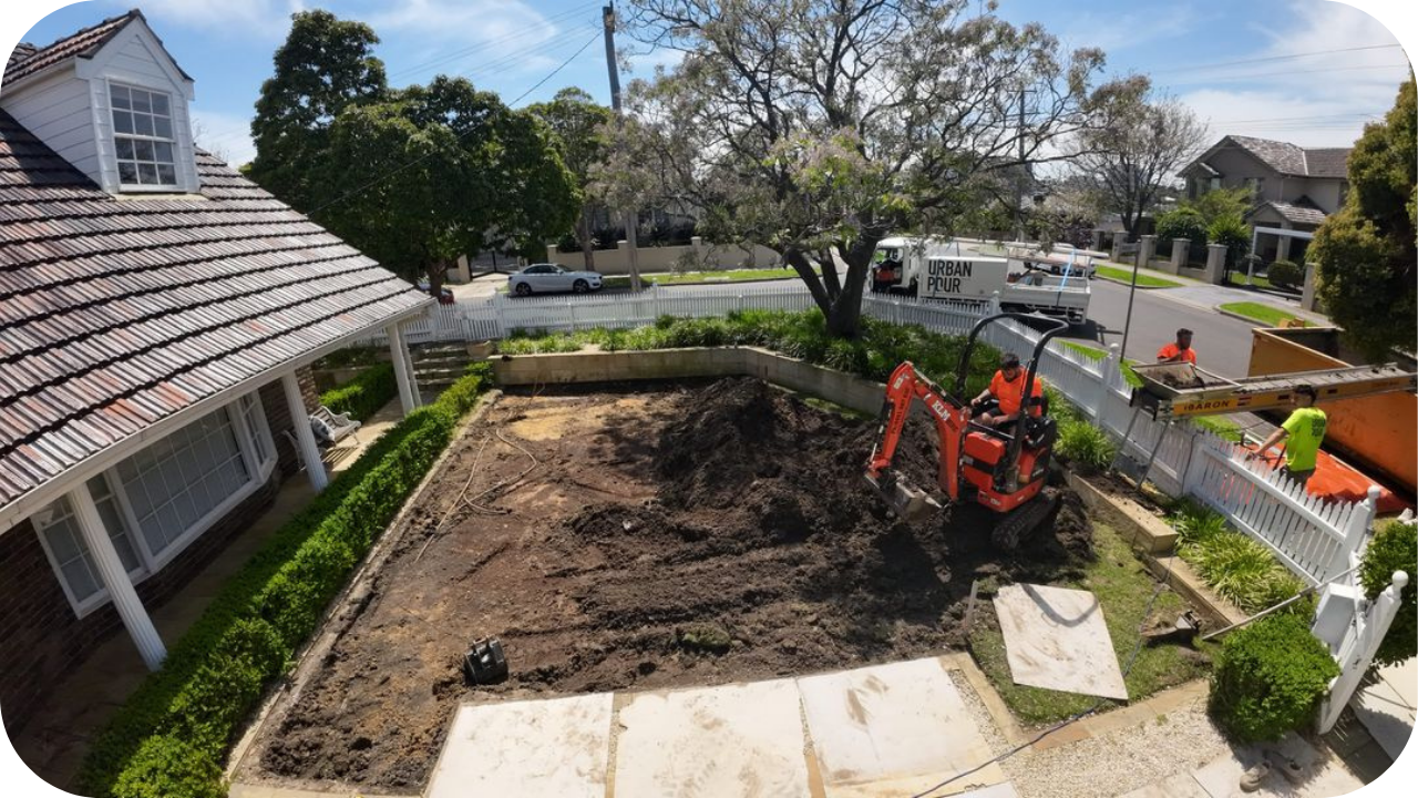 Construction crew operating a small excavator to clear and level soil in a residential front yard, with workers preparing the site near a white picket fence and an Urban Pour vehicle parked on the street.
