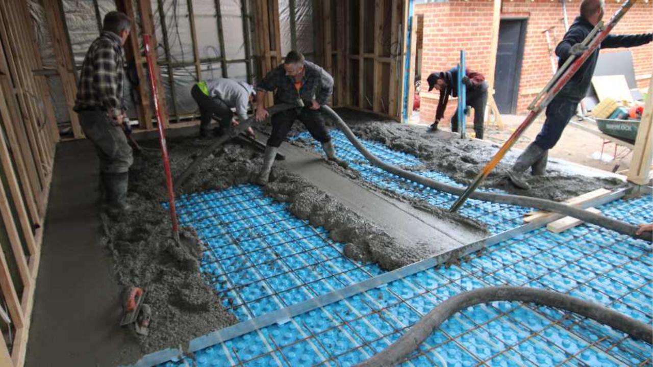 Crew spreading pumped concrete across a formwork layout with blue insulation panels and steel reinforcement during a controlled indoor slab pour