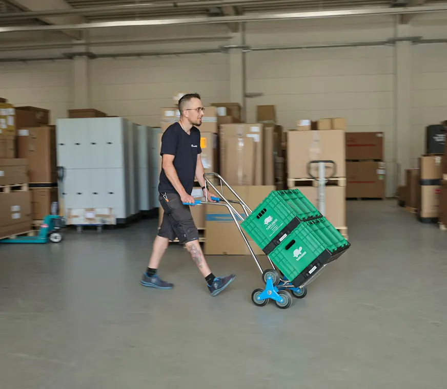 Grüner Umzug bei Pohl - ein Mitarbeiter Schiebt einen Wagen mit grünen Boxen durch ein Lager mit Kartons 