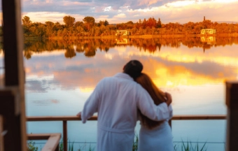 Couple in white robes embracing while overlooking a lake at sunset with autumn trees reflected in water.