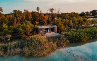 Modern wooden cabin on stilts surrounded by greenery beside a calm river under a blue sky.