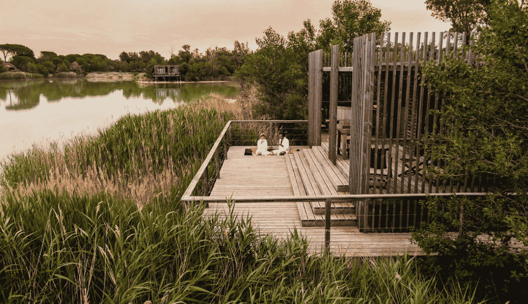 Two people sitting on a wooden deck by a lake surrounded by tall reeds and trees at sunset.