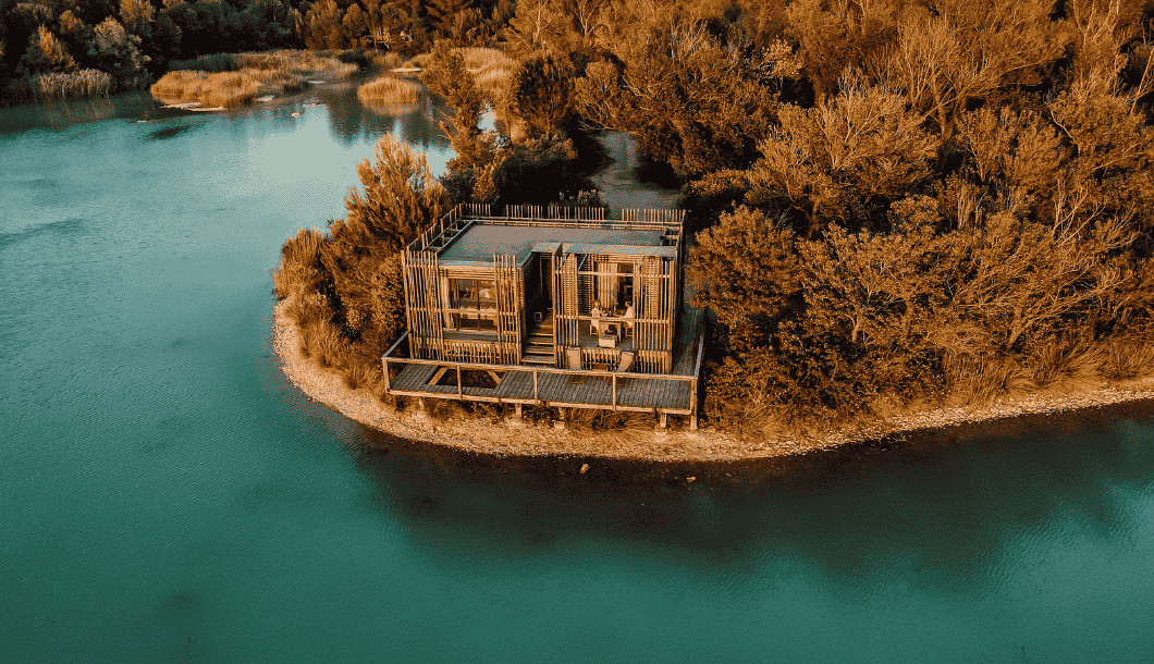 Modern wooden house with large windows on a small peninsula surrounded by turquoise water and autumn trees.