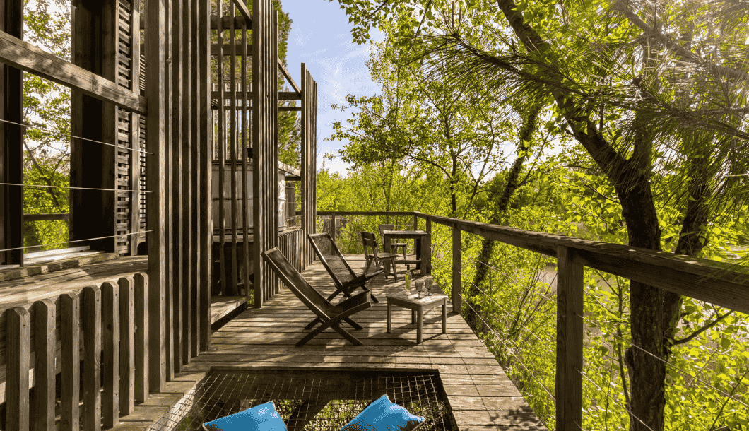 Wooden balcony with chairs, tables, and green trees bathed in sunlight.