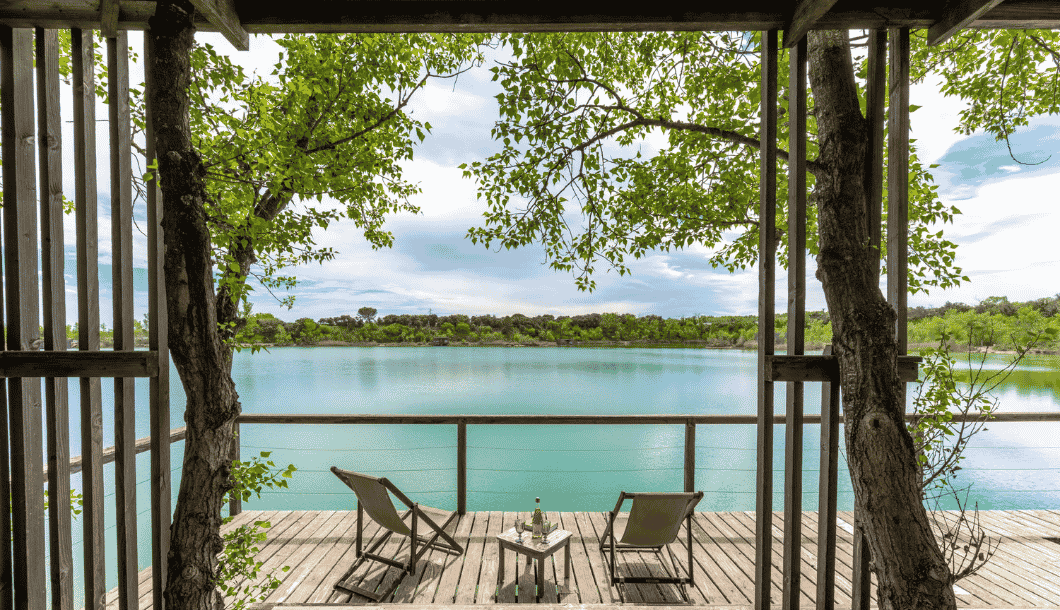 Deck with two chairs and a table overlooking a calm lake framed by green trees under a cloudy sky.