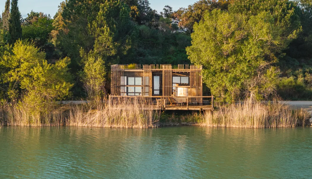 Wooden cabin with deck by a lake surrounded by tall grass and green trees during daylight.