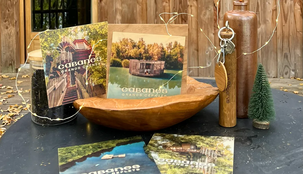 Wooden bowl on table holds two postcards with cabin images and a jar, keys, and small tree nearby.